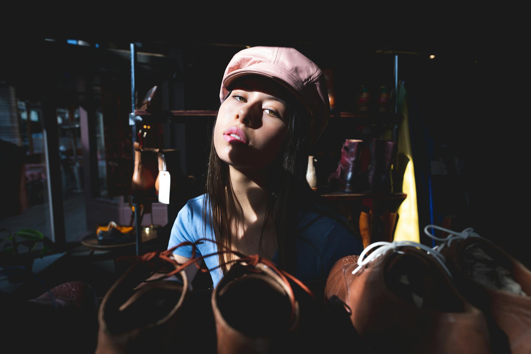 Chic woman posing with a trendy hat amidst shoes in a stylish setting, enhanced by dramatic sunlight.
